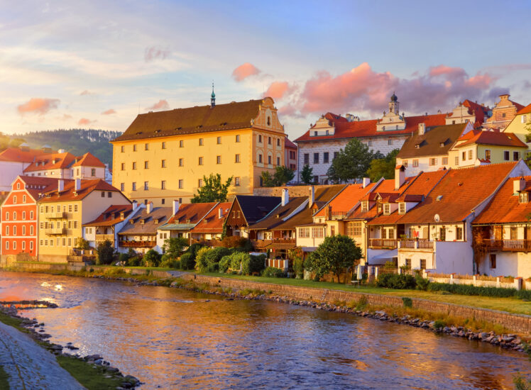 Český Krumlov při západu slunce, pohled od řeky Vltavy na historické domy a zámek v teplém večerním světle.