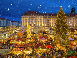 Sníh nad večerním Striezelmarktem v Drážďanech s vánoční výzdobou a davem návštěvníků.