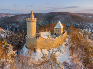 Zimní letecký pohled na hrad Kokořín pokrytý sněhem, obklopený lesy Kokořínska při západu slunce.