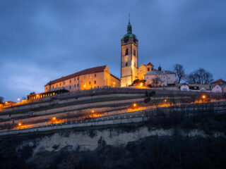 Osvětlený zámek Mělník a vinice při modré hodině nad soutokem Labe a Vltavy.