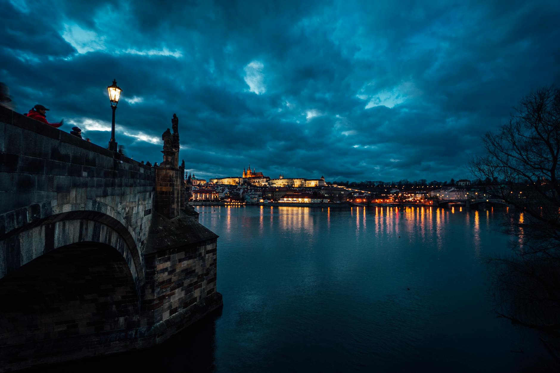 Karlův most v&nbsp;Praze v&nbsp;modré hodině — Pražský hrad se rozsvěcuje a&nbsp;Vltava zrcadlí dramatickou oblohu