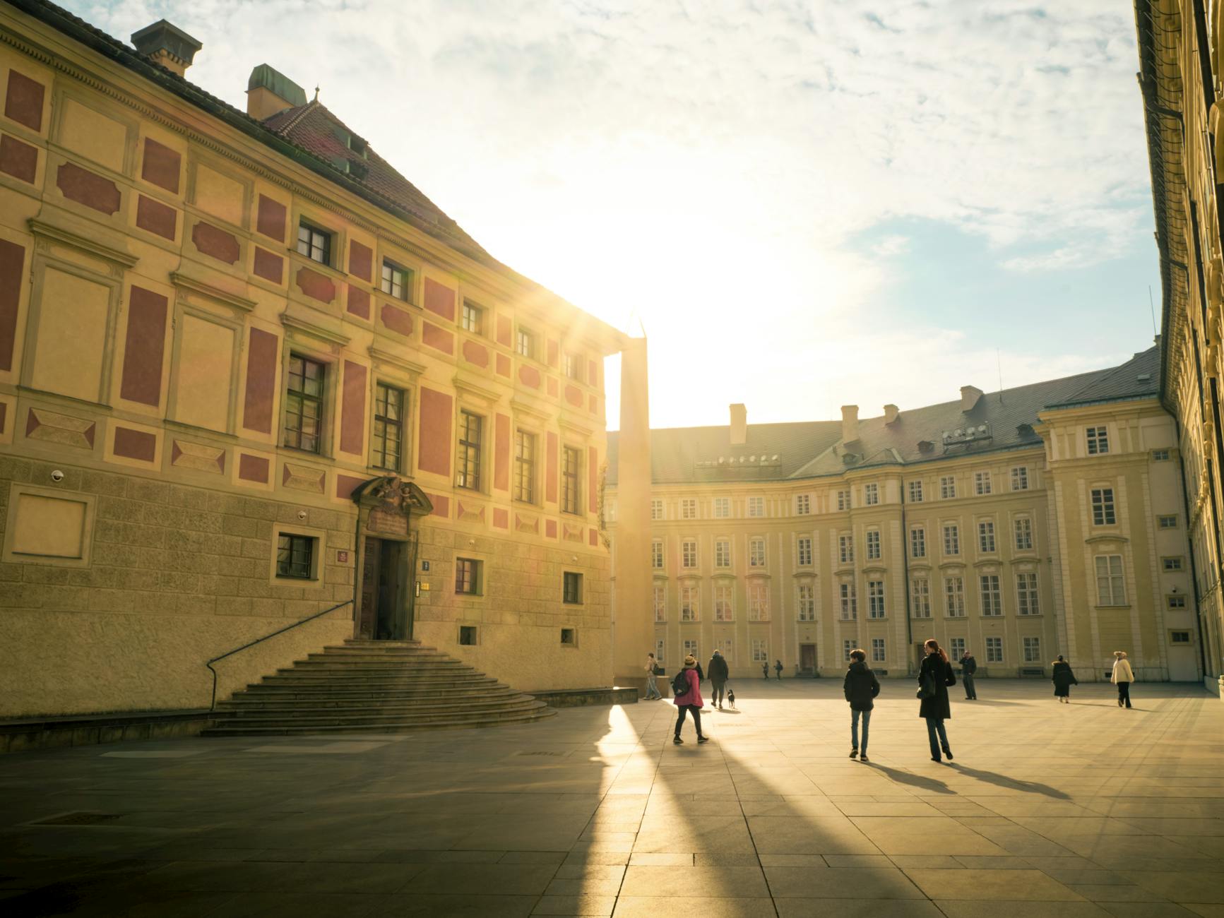 A sunlit courtyard featuring historical European architecture and people walking, casting long shadows.